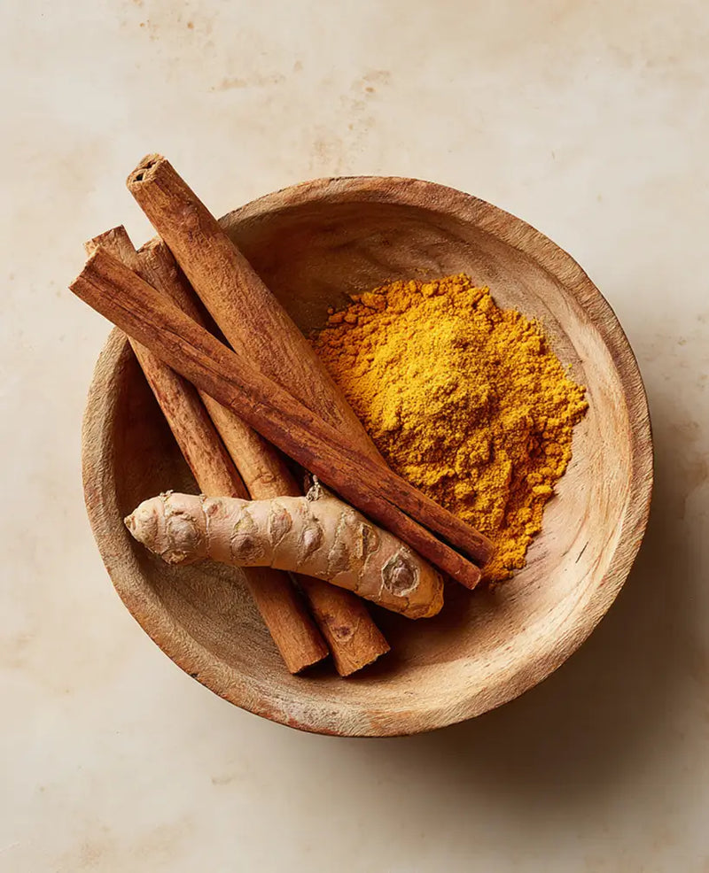 Wooden bowl with turmeric powder, cinnamon sticks, and a piece of ginger on a beige background